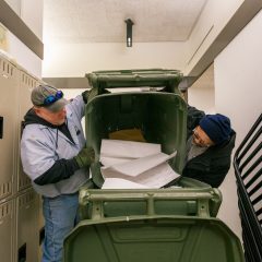 Two building service workers emptying a large recycling bin of paper