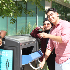 Two people placing something into a Big Belly recycling bin